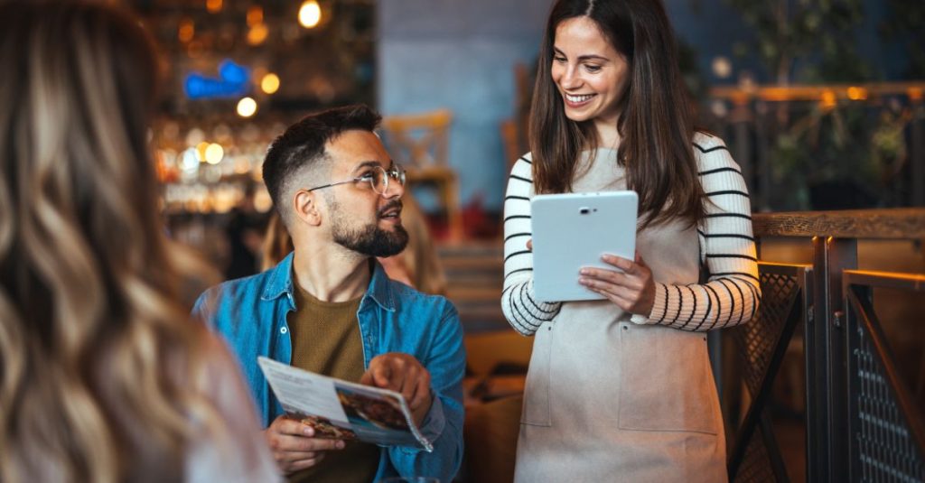 A cheerful waitress holding a tablet takes an order from a seated male customer holding a menu in a warmly lit restaurant, both smiling and engaged in friendly conversation.