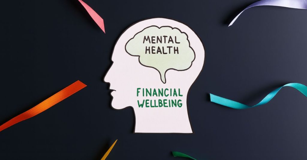 A paper cutout of a human head silhouette on a dark blue background. Inside the head, a green brain shape contains the words “MENTAL HEALTH,” while the lower part of the head reads “FINANCIAL WELLBEING” in green text. Colourful ribbons in red, orange, yellow, blue, and purple radiate outward, symbolising the connection between mental and financial wellbeing.